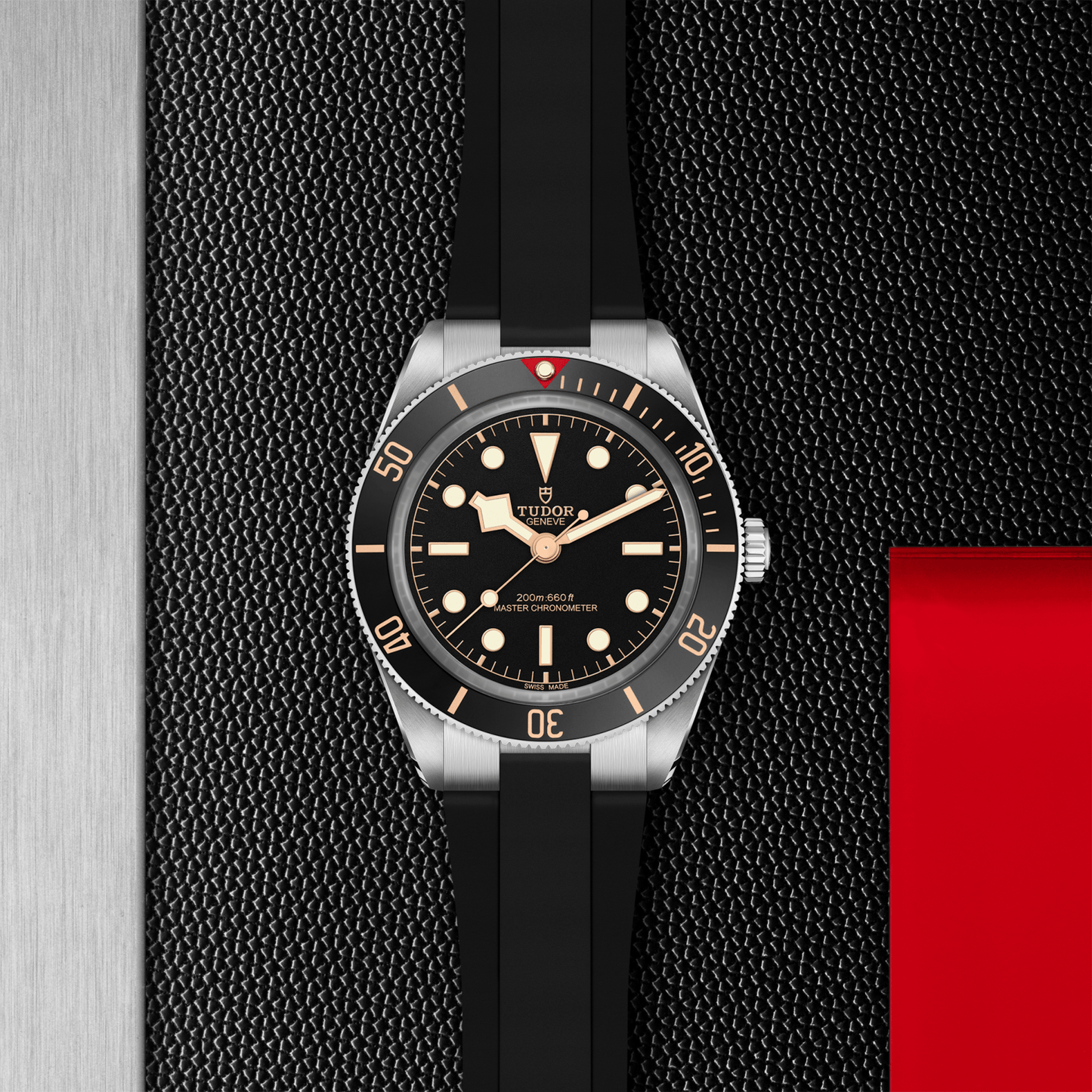 The TUDOR Black Bay 58 dive watch features a black bezel and dial, luminous hour markers and hands, and comes on a rubber strap. It sits on a textured black surface accented with silver and red highlights.