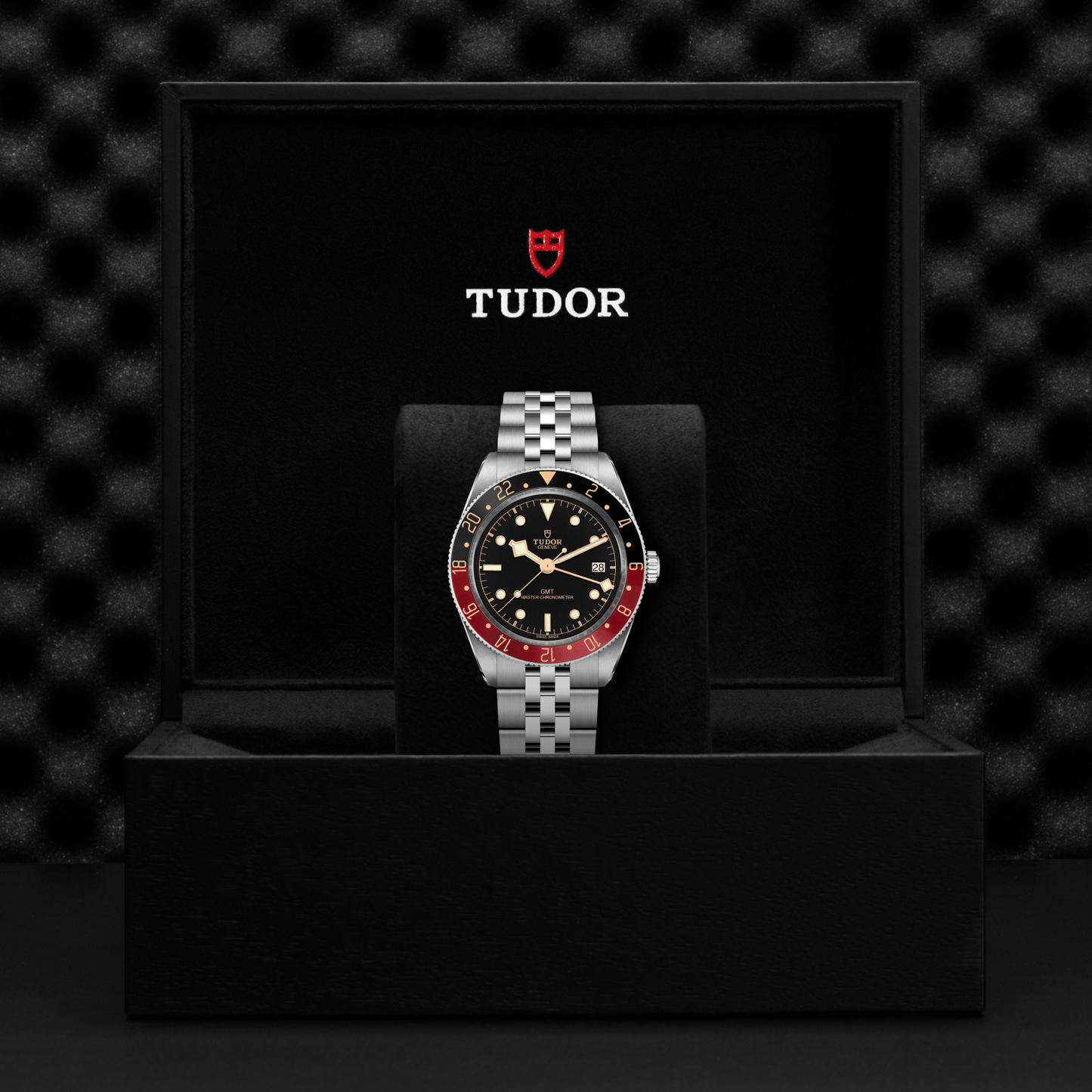A close-up of the TUDOR Black Bay 58 GMT wristwatch, featuring a black dial and red-black bezel, shown in an open black TUDOR box with cushioned interior and logo inside the lid. Powered by the GMT Manufacture Caliber.