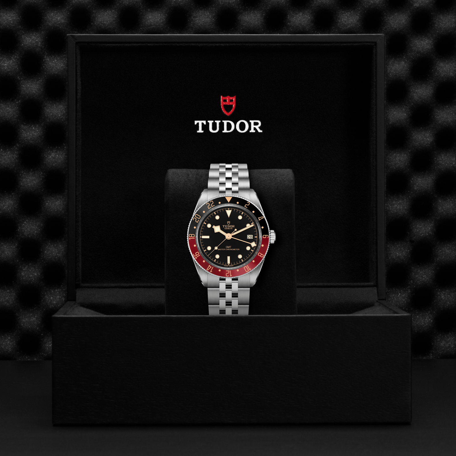A close-up of the TUDOR Black Bay 58 GMT wristwatch, featuring a black dial and red-black bezel, shown in an open black TUDOR box with cushioned interior and logo inside the lid. Powered by the GMT Manufacture Caliber.