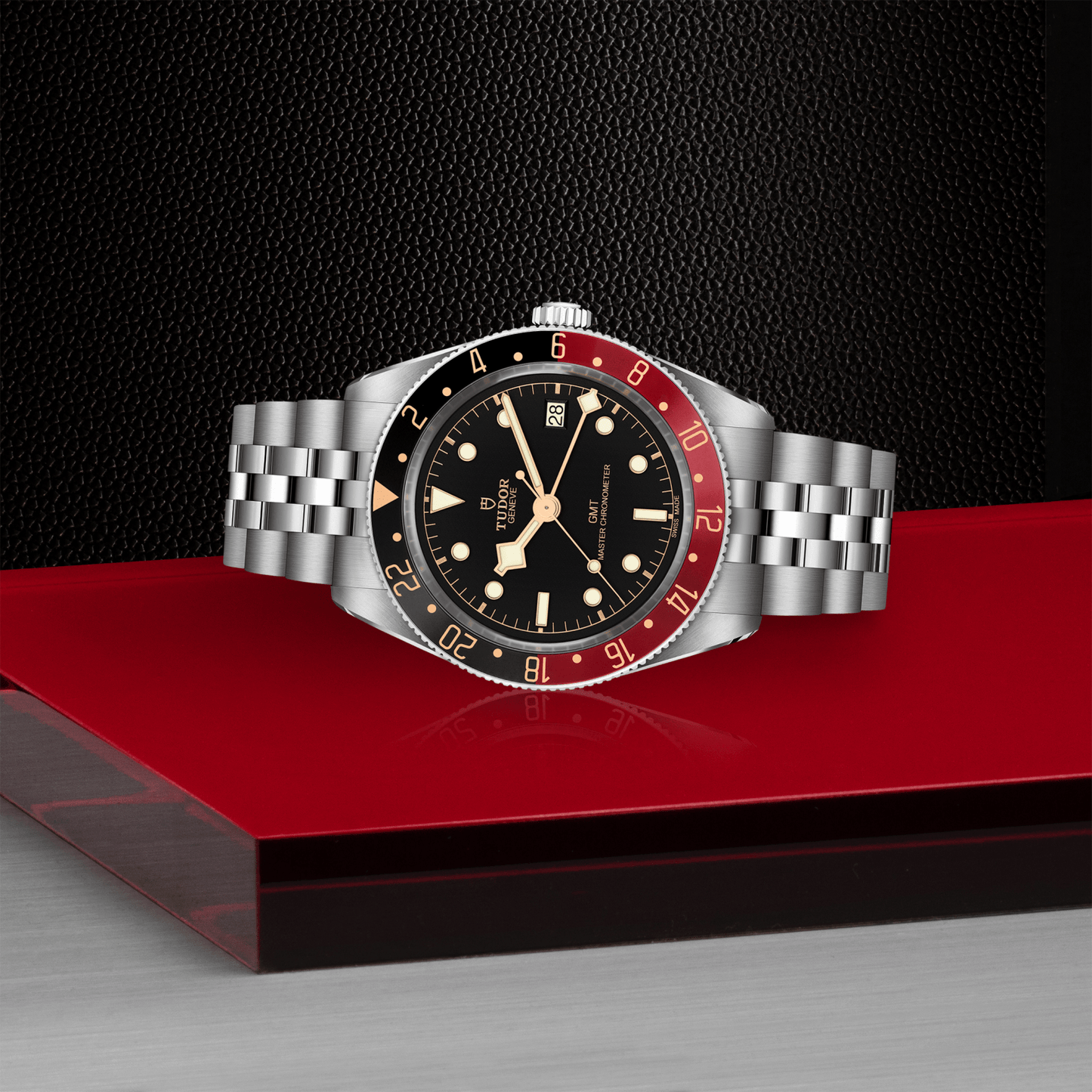 The TUDOR Black Bay 58 GMT wristwatch, featuring a black and red bezel, black dial, and silver bracelet, rests on a glossy red surface with a textured black background. It showcases the advanced TUDOR GMT Manufacture Caliber.