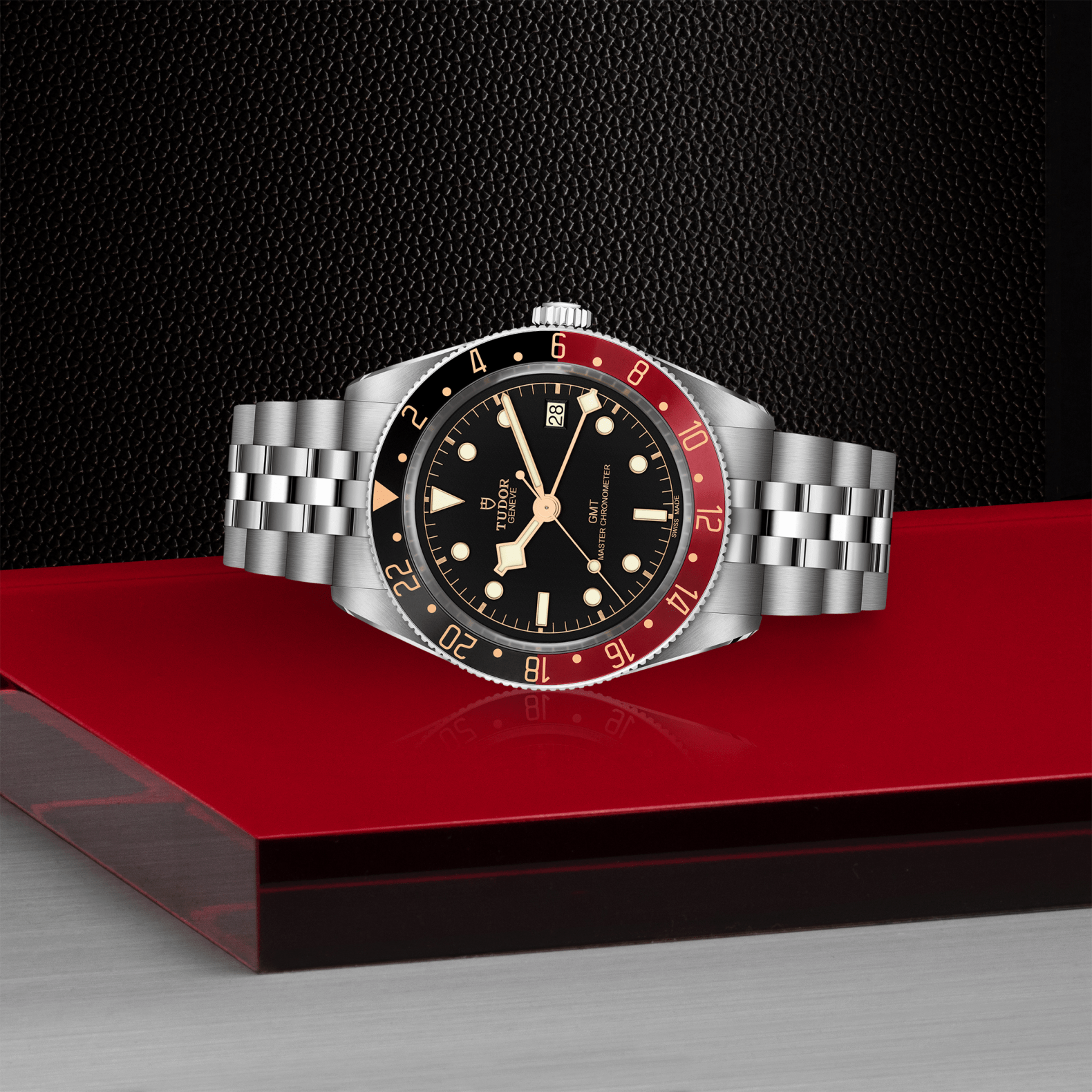 The TUDOR Black Bay 58 GMT wristwatch, featuring a black and red bezel, black dial, and silver bracelet, rests on a glossy red surface with a textured black background. It showcases the advanced TUDOR GMT Manufacture Caliber.