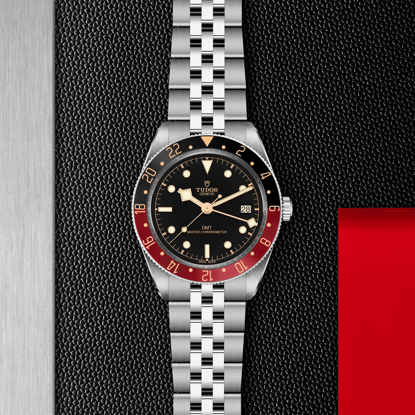 A TUDOR Black Bay 58 GMT wristwatch by TUDOR with a silver metal bracelet rests on a textured black surface, showcasing a black dial, red and black bezel, gold accents, the GMT Manufacture Caliber, and a date window at 3 o’clock.
