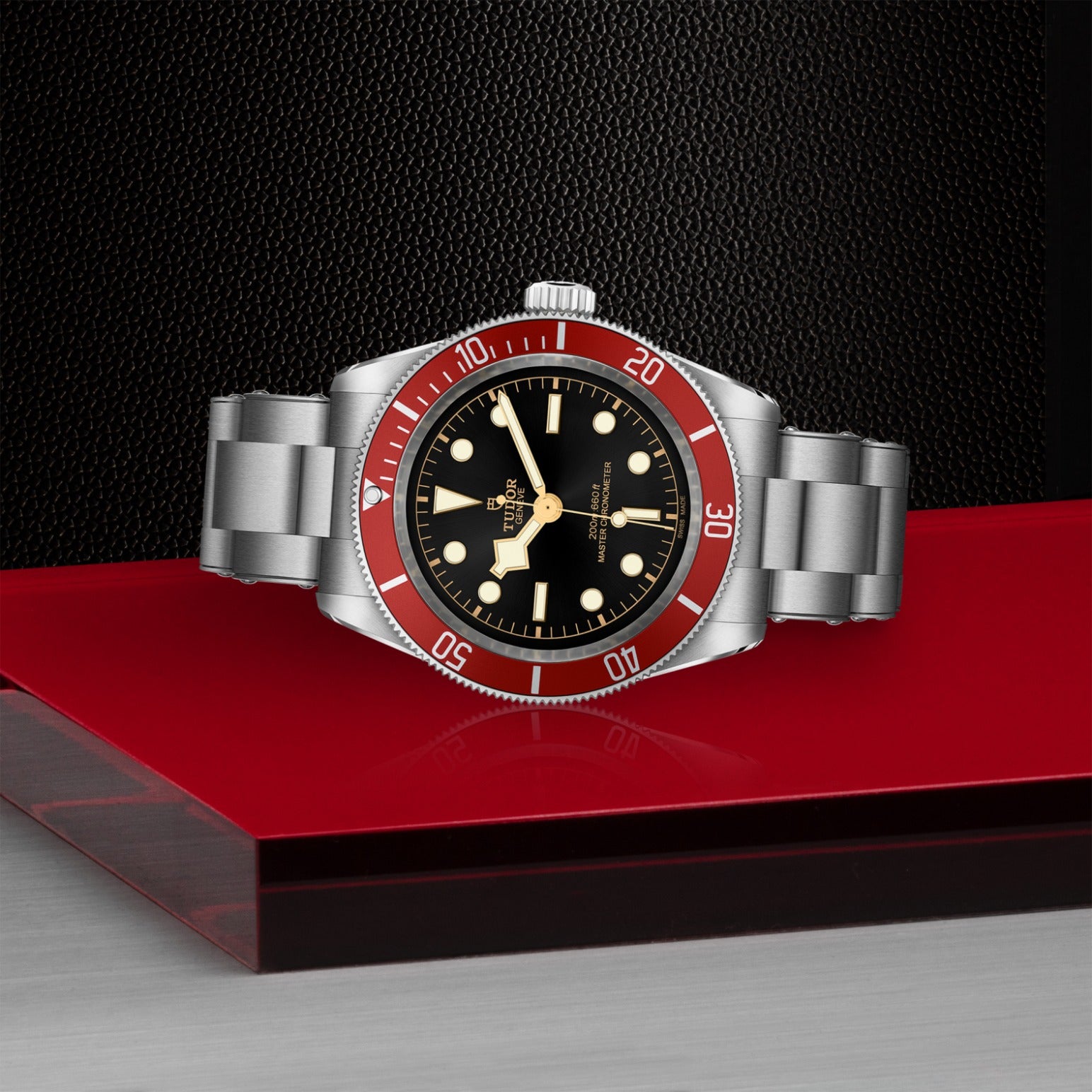 The TUDOR Black Bay by TUDOR features a 41mm steel case, stainless steel band, black dial with bold white markers, red bezel, and gold accents—shown on a red and metallic surface set against a textured black background.