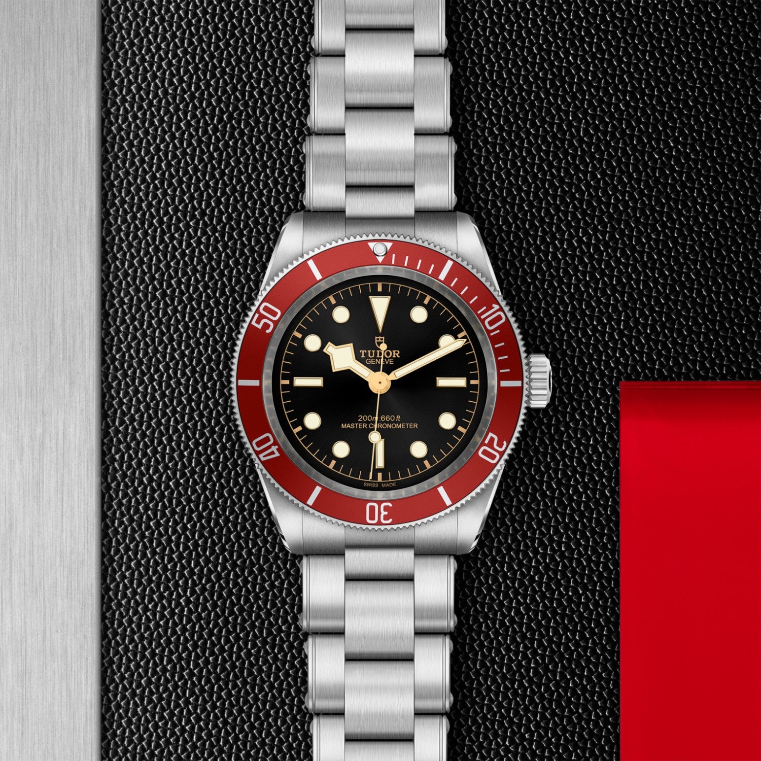 A TUDOR Black Bay wristwatch with a stainless steel bracelet and red bezel rests on a black surface beside silver and red strips. It features a 41mm steel case, black dial, and signature gold accents from TUDOR.