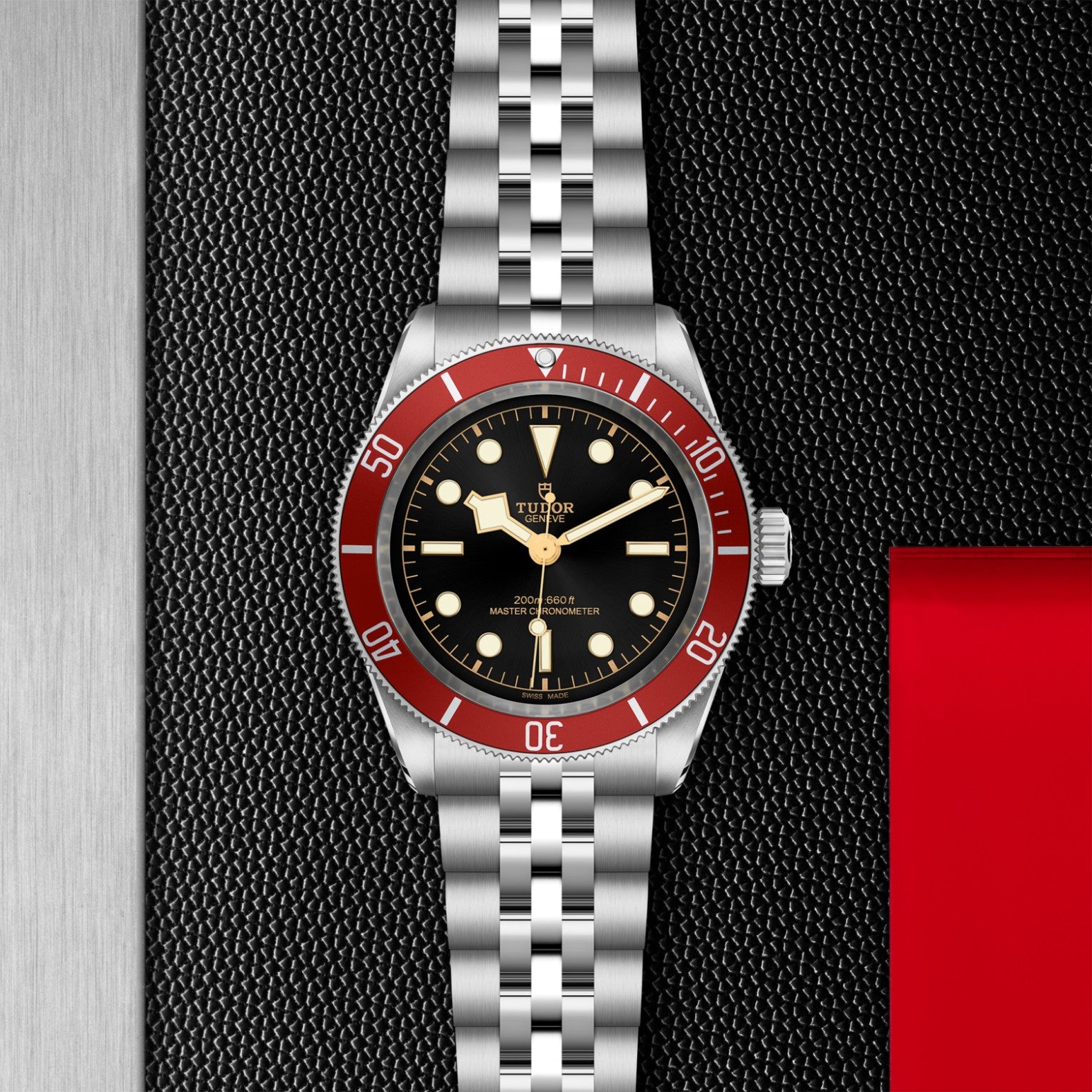 The TUDOR Black Bay by TUDOR, a neo-vintage stainless steel wristwatch with a red bezel, black dial, and silver jubilee bracelet, is displayed vertically on a textured black background with red and metallic silver accents.