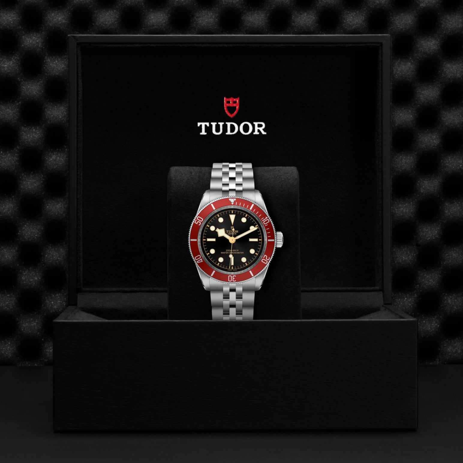 A TUDOR Black Bay wristwatch with a silver metal band, black dial, and red bezel is displayed upright in an open black TUDOR box with the brand logo visible inside the lid.