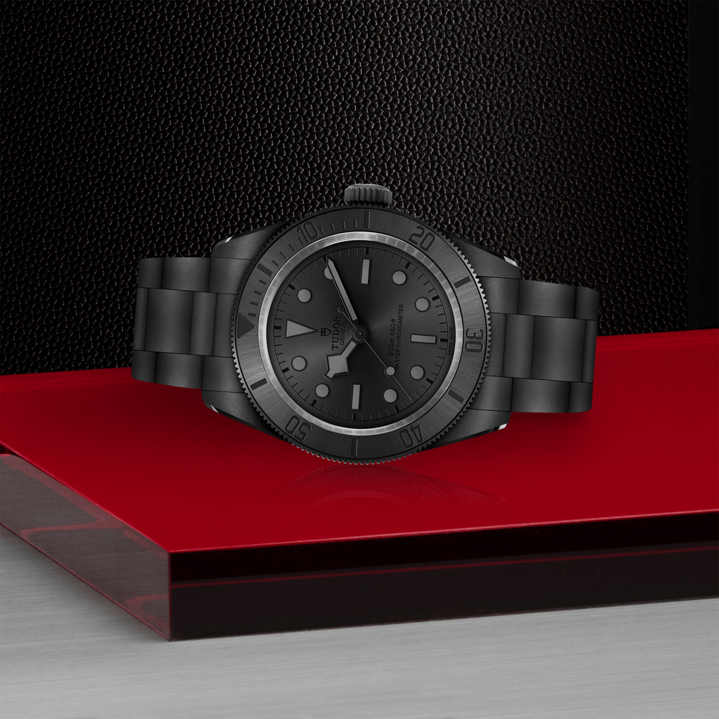 A TUDOR Black Bay Ceramic matte black wristwatch sits on a glossy red platform with a metal band, contrasting against a textured black background and metallic surface.