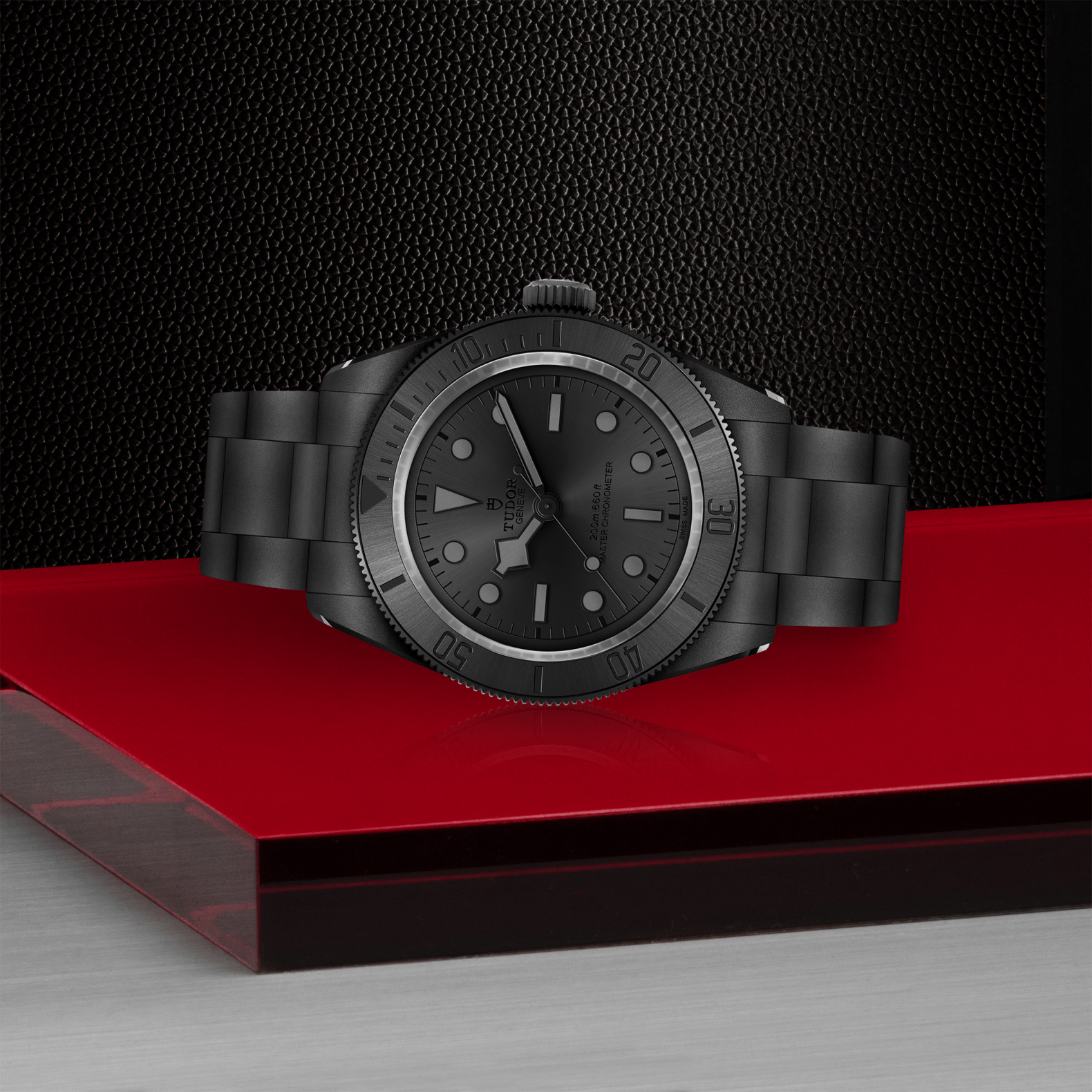 A TUDOR Black Bay Ceramic matte black wristwatch sits on a glossy red platform with a metal band, contrasting against a textured black background and metallic surface.