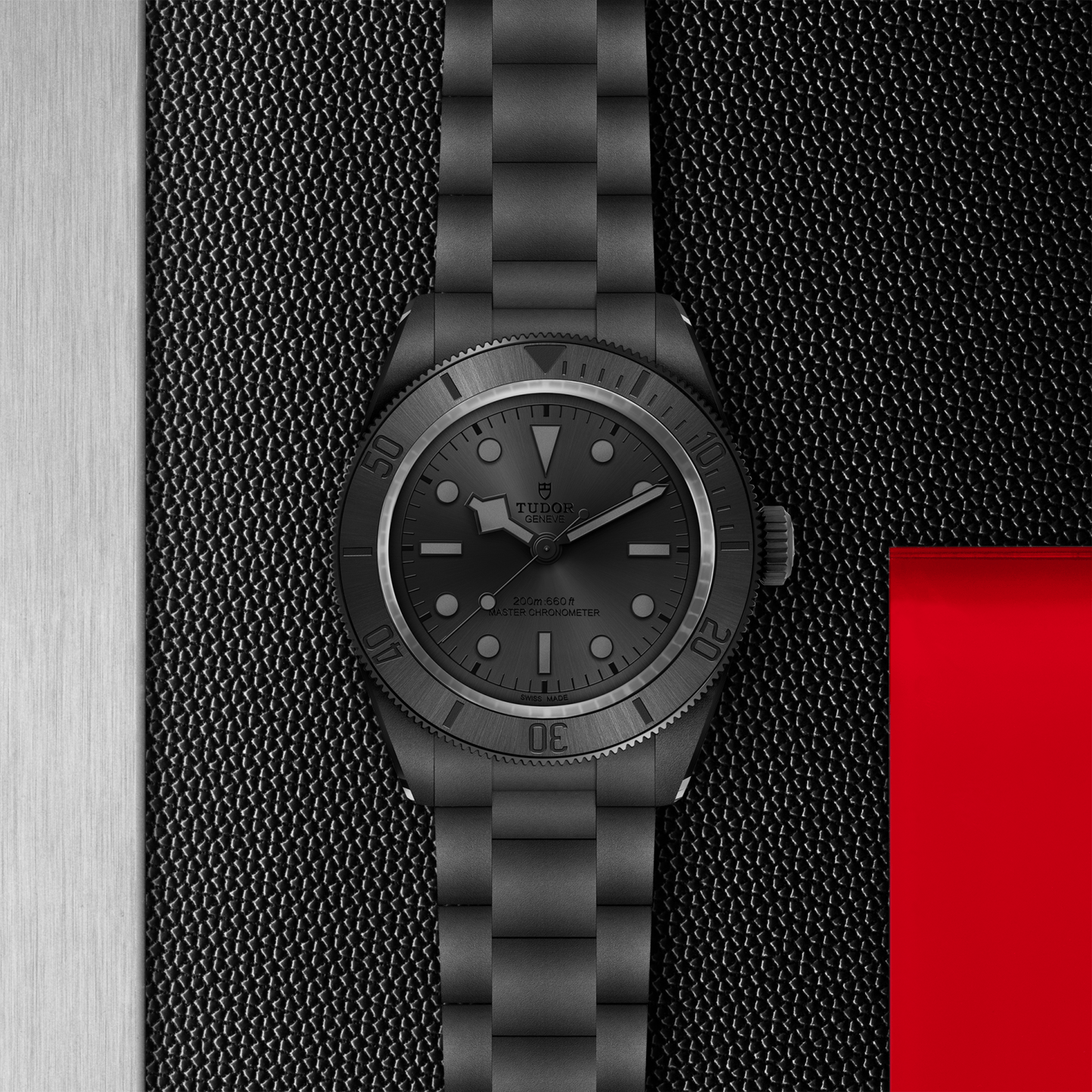 A TUDOR Black Bay Ceramic wristwatch in matte black with a metal bracelet sits on a textured black surface, bordered by brushed steel on the left and a red rectangle at the lower right.