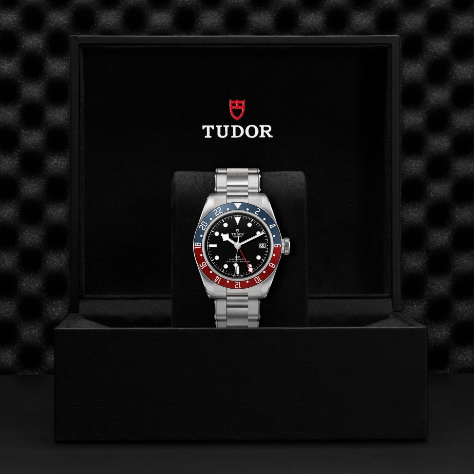 A TUDOR Black Bay GMT wristwatch with a steel case and red-blue bezel is shown in an open black TUDOR box, the logo visible inside the lid. The watch features multiple time zones and rests against a dark, textured background.