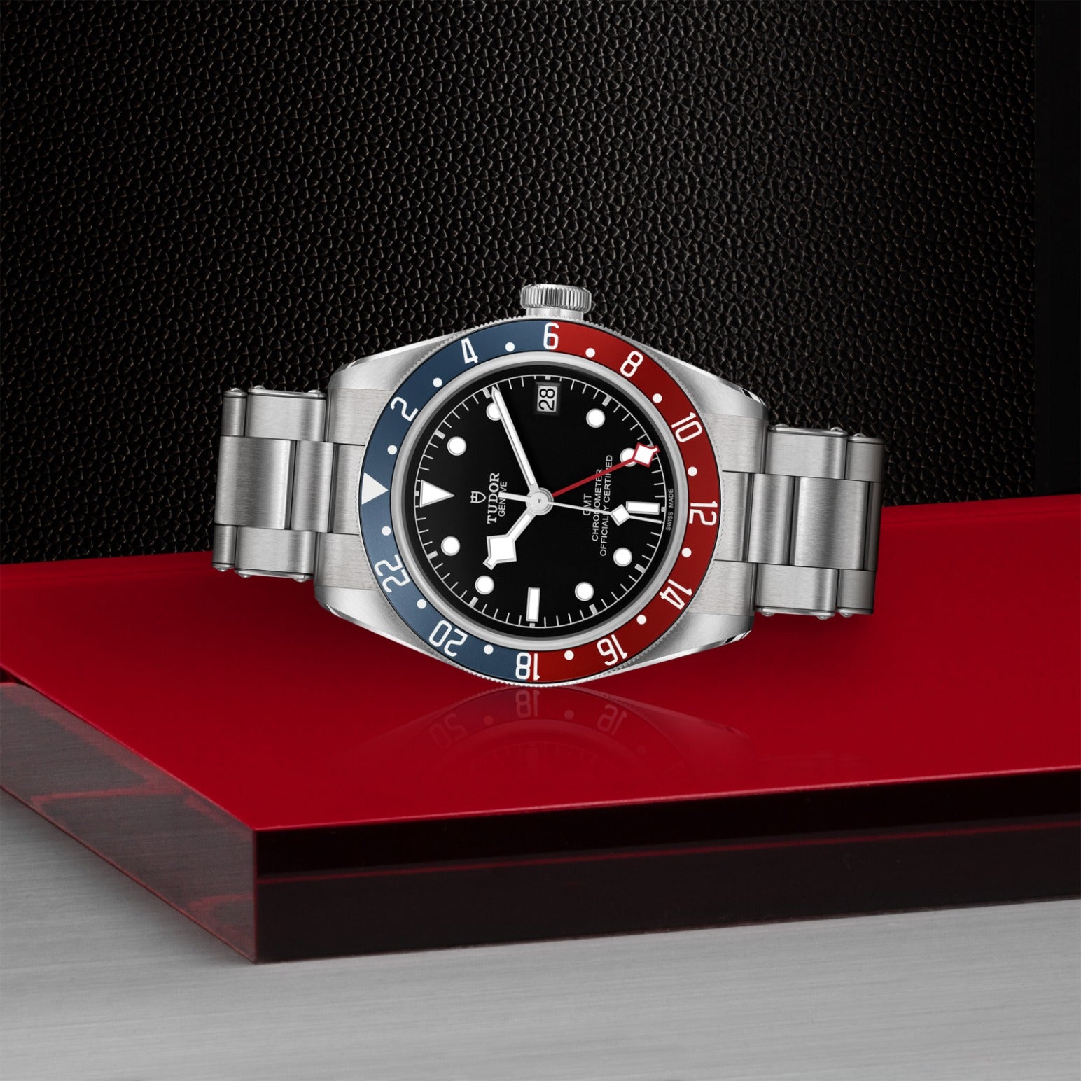 The TUDOR Black Bay GMT wristwatch, featuring a steel case, black dial, silver markers, and a blue-and-red bezel, rests on a glossy red surface with a textured black background.