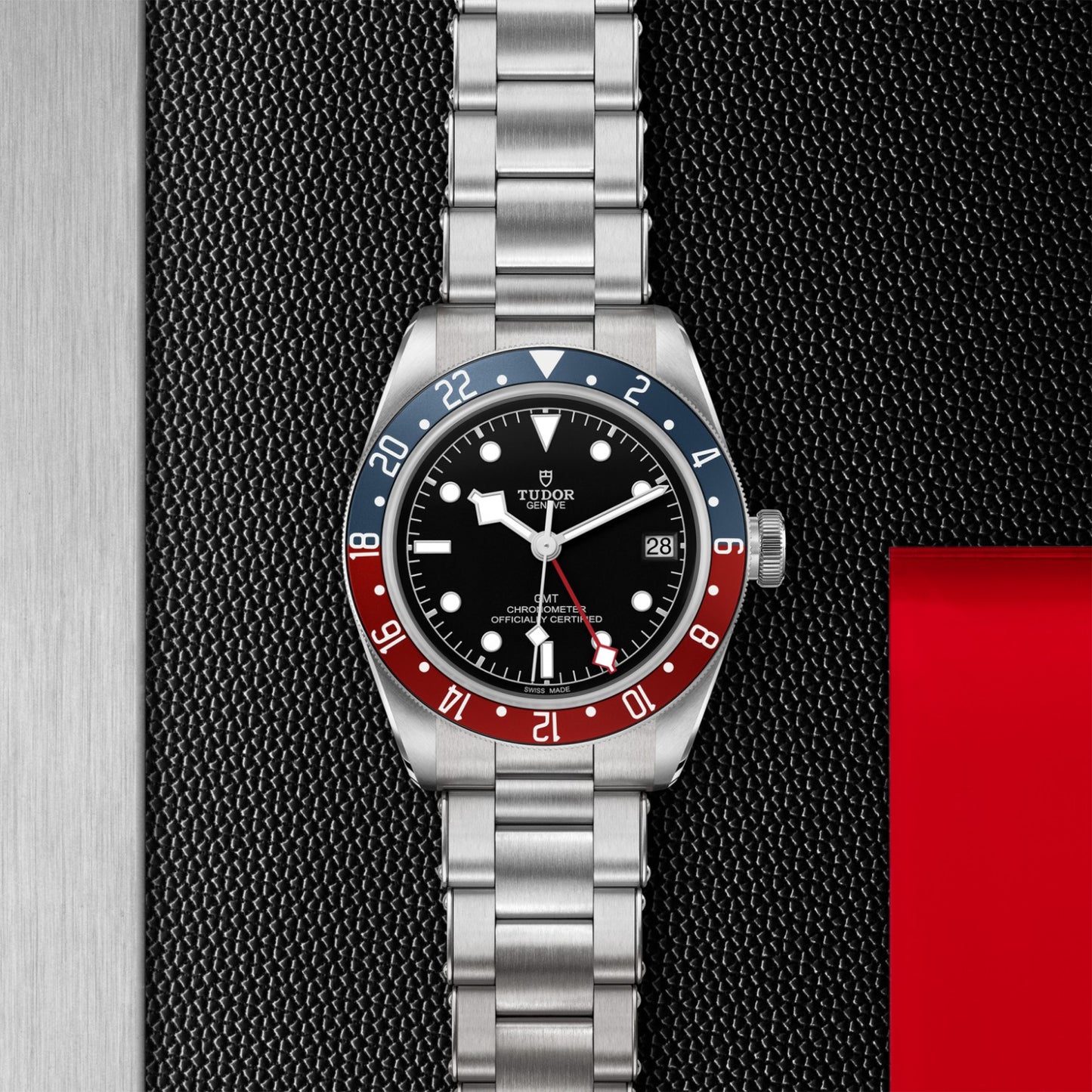 The TUDOR Black Bay GMT wristwatch features a stainless steel case, black dial, date display, and a blue and red bezel. Displayed on a textured black and metal background with a red accent in the corner. Brand: TUDOR.