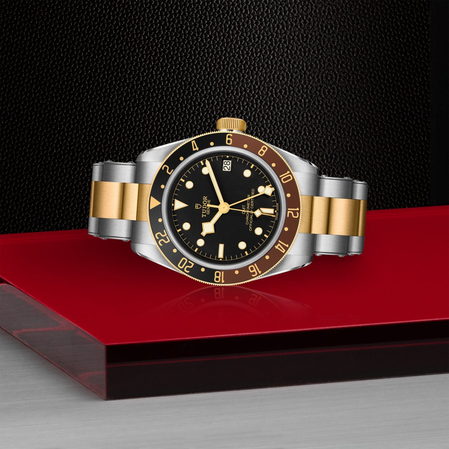 The TUDOR Black Bay GMT S&G wristwatch, featuring a black dial, gold and silver bracelet, rotatable bezel, and date display, is showcased on a reflective red surface with a textured black background.