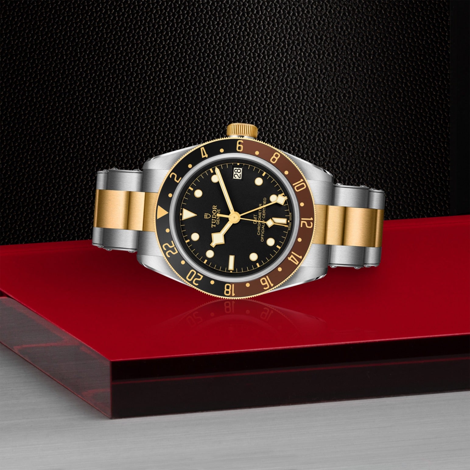 The TUDOR Black Bay GMT S&G wristwatch, featuring a black dial, gold and silver bracelet, rotatable bezel, and date display, is showcased on a reflective red surface with a textured black background.
