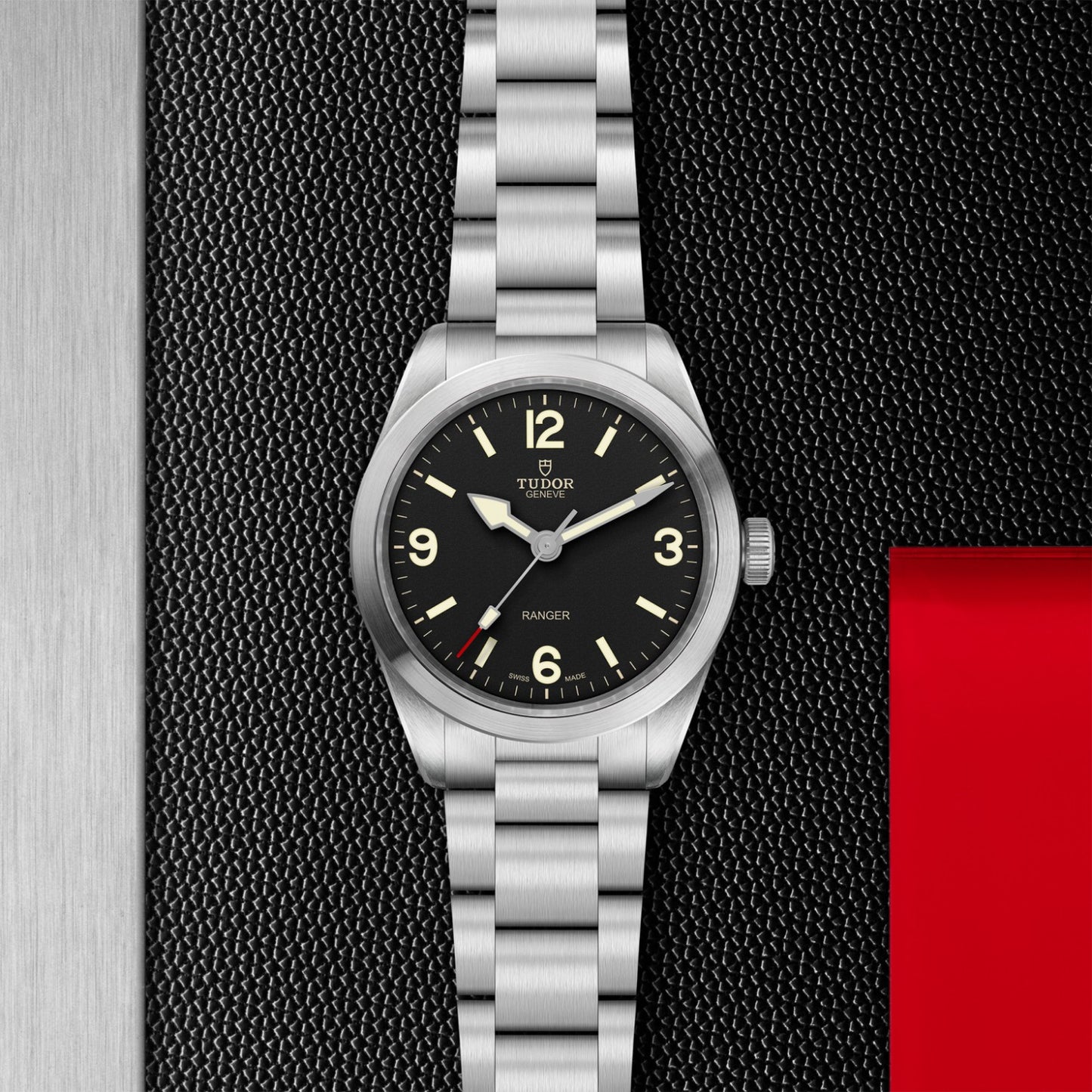The TUDOR Ranger, featuring a 39mm steel case, stainless steel bracelet, black dial, and bold numerals at 12, 3, 6, and 9 with luminous markers, is showcased on a textured black and red background.