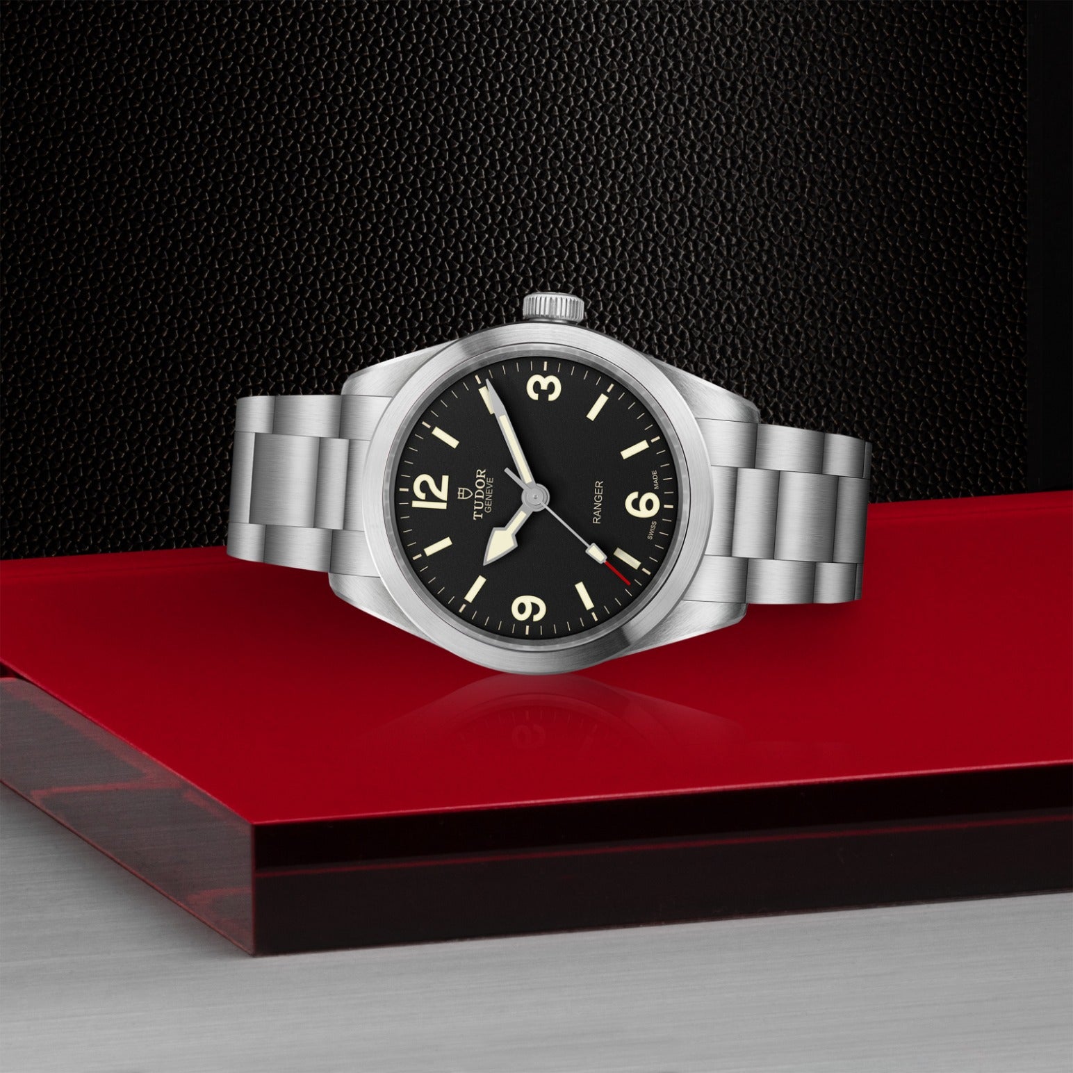 The TUDOR Ranger watch by TUDOR features a 39mm steel case, black dial, and luminescent markers, displayed on a shiny red surface with a textured black background and metallic accents.