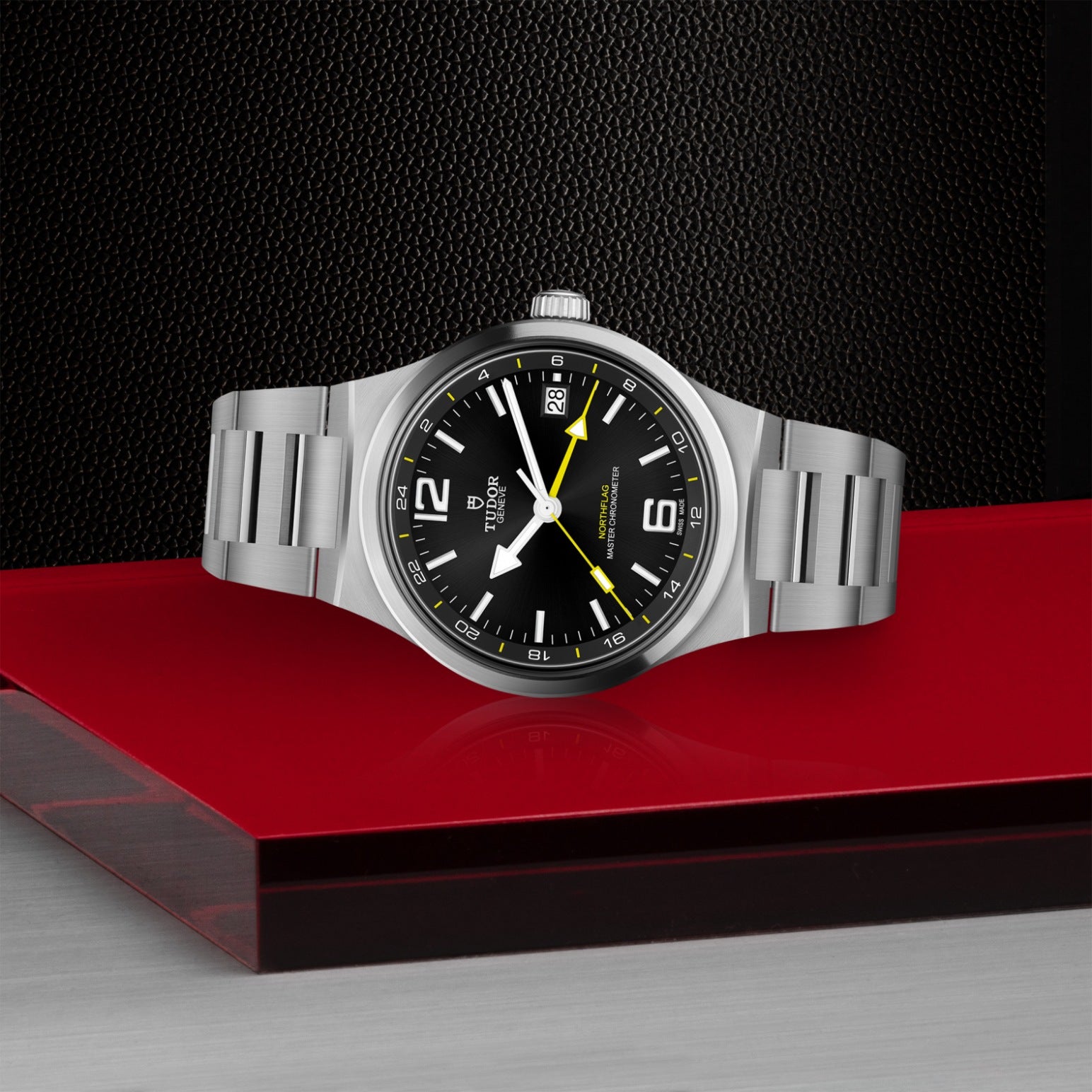 A TUDOR North Flag wristwatch by TUDOR, featuring a stainless steel case, black dial with yellow accents, and date display, is showcased on a glossy red platform against a textured black and silver backdrop.