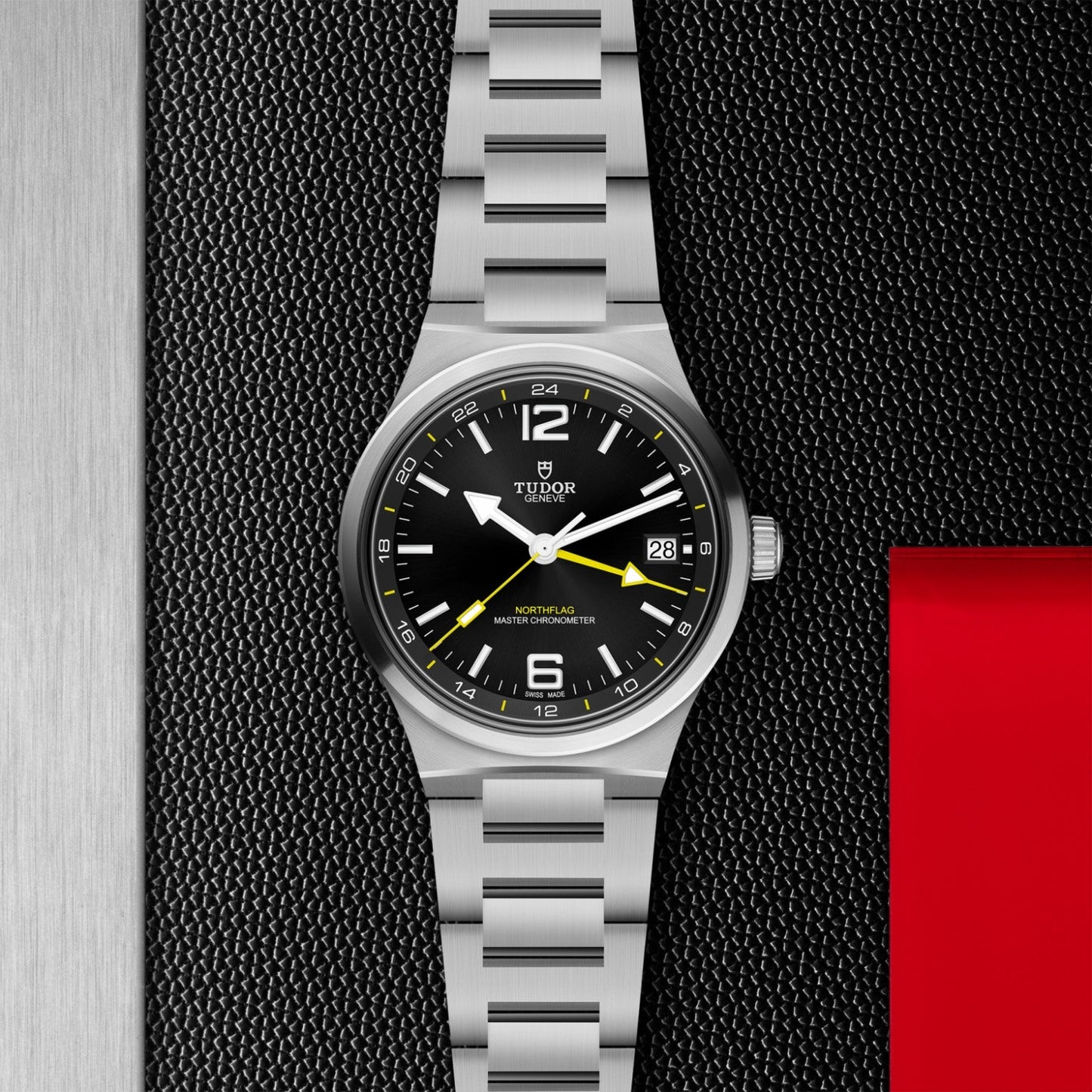 A TUDOR North Flag watch in silver, featuring a black dial with yellow accents and a metallic band, is displayed on a textured black surface beside a red and silver background. The time on the watch is set to 10:10.