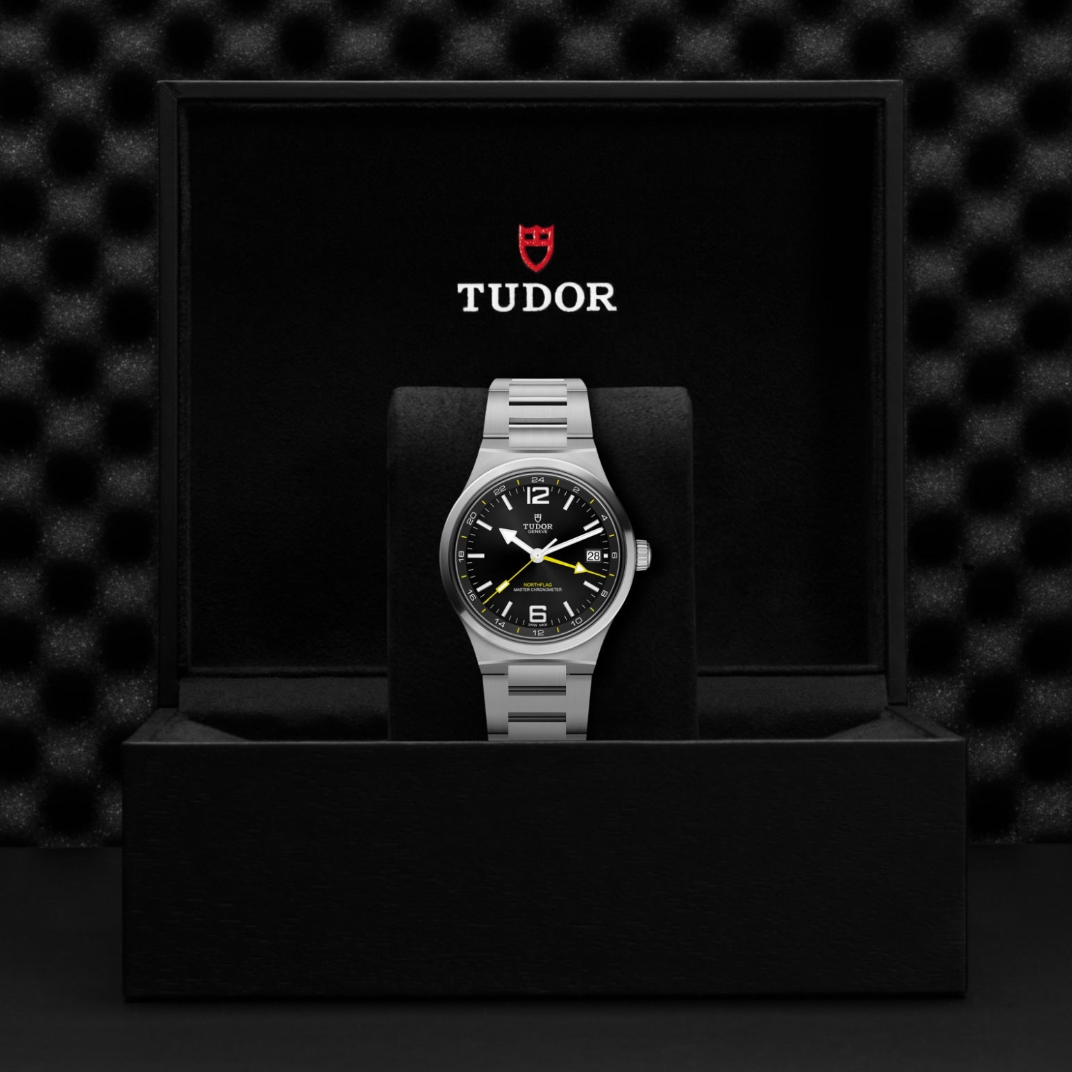 A TUDOR North Flag wristwatch with a silver band is displayed in an open black box featuring the TUDOR logo and name above it. With Master Chronometer certification, the watch showcases a black face with white and yellow details inspired by the iconic TUDOR North Flag design. The background is dark and textured.