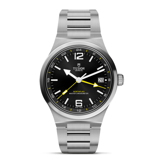 A TUDOR North Flag stainless steel wristwatch featuring a black dial, yellow GMT hand, date window at 3 o’clock, silver-tone indices, and a brushed metal bracelet exemplifies the brand’s spirit of precision—all presented against a white backdrop.