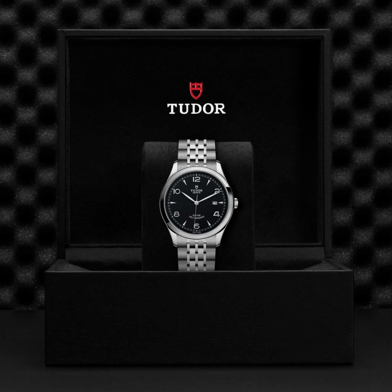 A silver TUDOR 1926 wristwatch with a black dial is showcased upright in an open black TUDOR box against a textured black background, highlighting the brand’s timeless elegance.