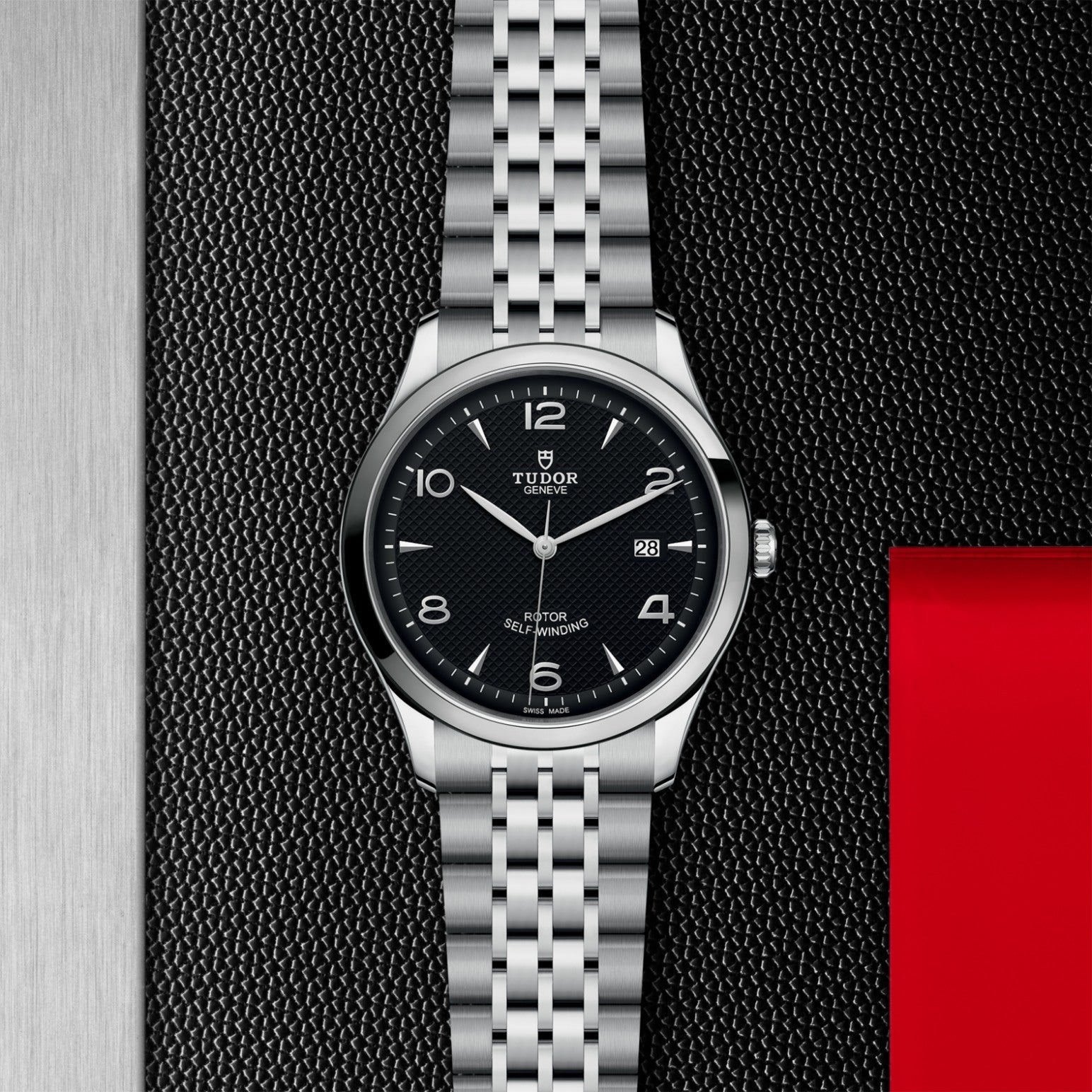 The TUDOR 1926, a classic silver wristwatch with a black dial, silver markers, date display, and metal link bracelet from TUDOR, is displayed on a textured dark surface with a silver and red border for an elegant look.