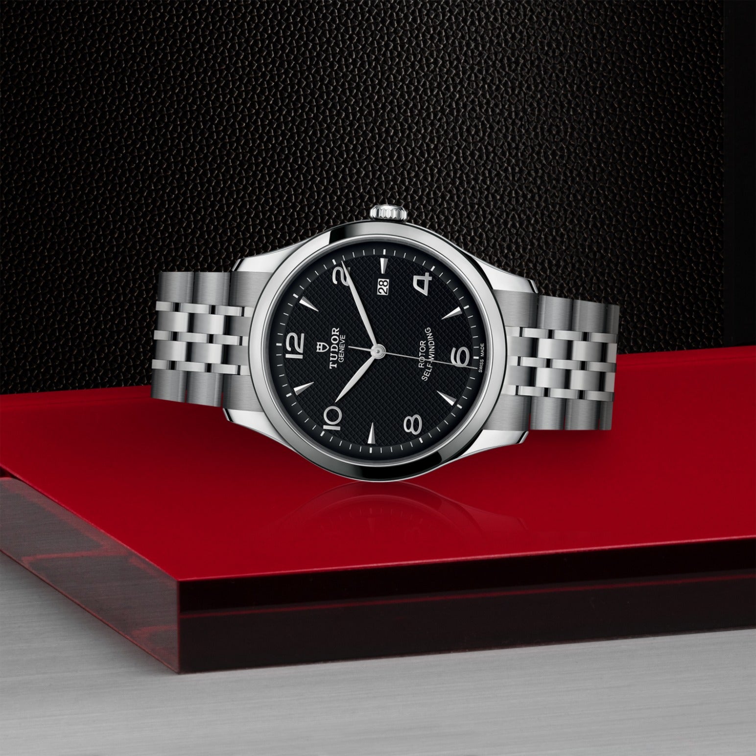 The TUDOR 1926, a silver wristwatch with black dial and metal link band by TUDOR, is shown on a red and gray surface against a textured black background, highlighting its classic elegance.