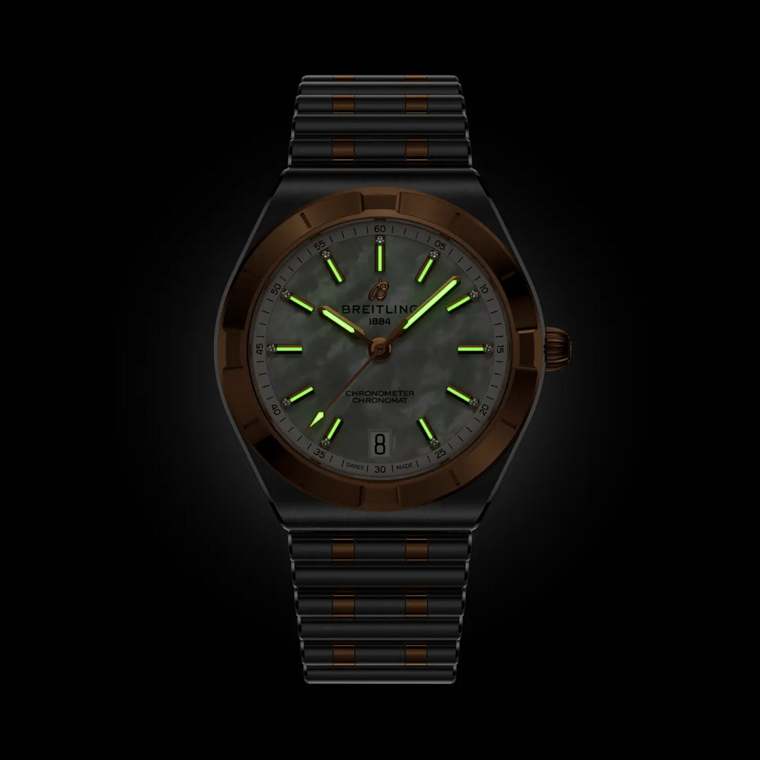 The BREITLING Chronomat Automatic 36 is a luxury sports watch with a dark metal band, brown accents, and a black mother-of-pearl dial. Its COSC-certified hands and markers glow green in the dark.