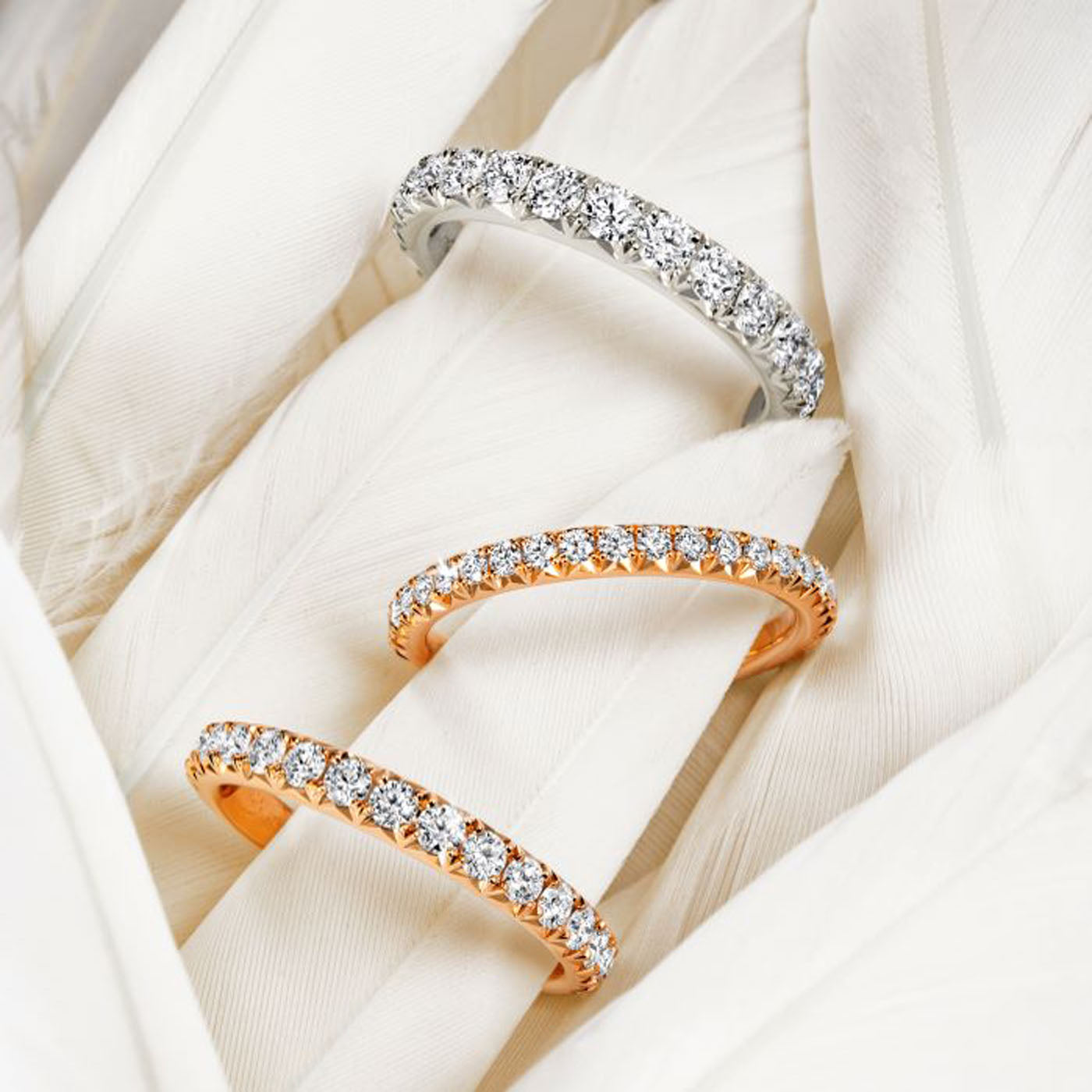 Three diamond-studded rings are elegantly placed on soft, white feathers. The top ring, a Hearts On Fire Vela Platinum Round Brilliant Diamond Anniversary Ring, sits above two rose gold Hearts On Fire Vela rings, creating a striking contrast against the delicate background.