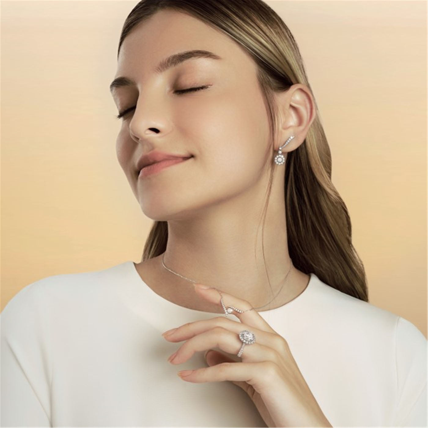A woman with long hair, dressed in a white shirt, elegantly models her jewelry. With her eyes softly closed, she showcases the exquisite Hearts On Fire Vela 18K White Gold Drop Earrings paired with a dazzling Hearts On Fire diamond ring. The background features a soft gradient of beige.