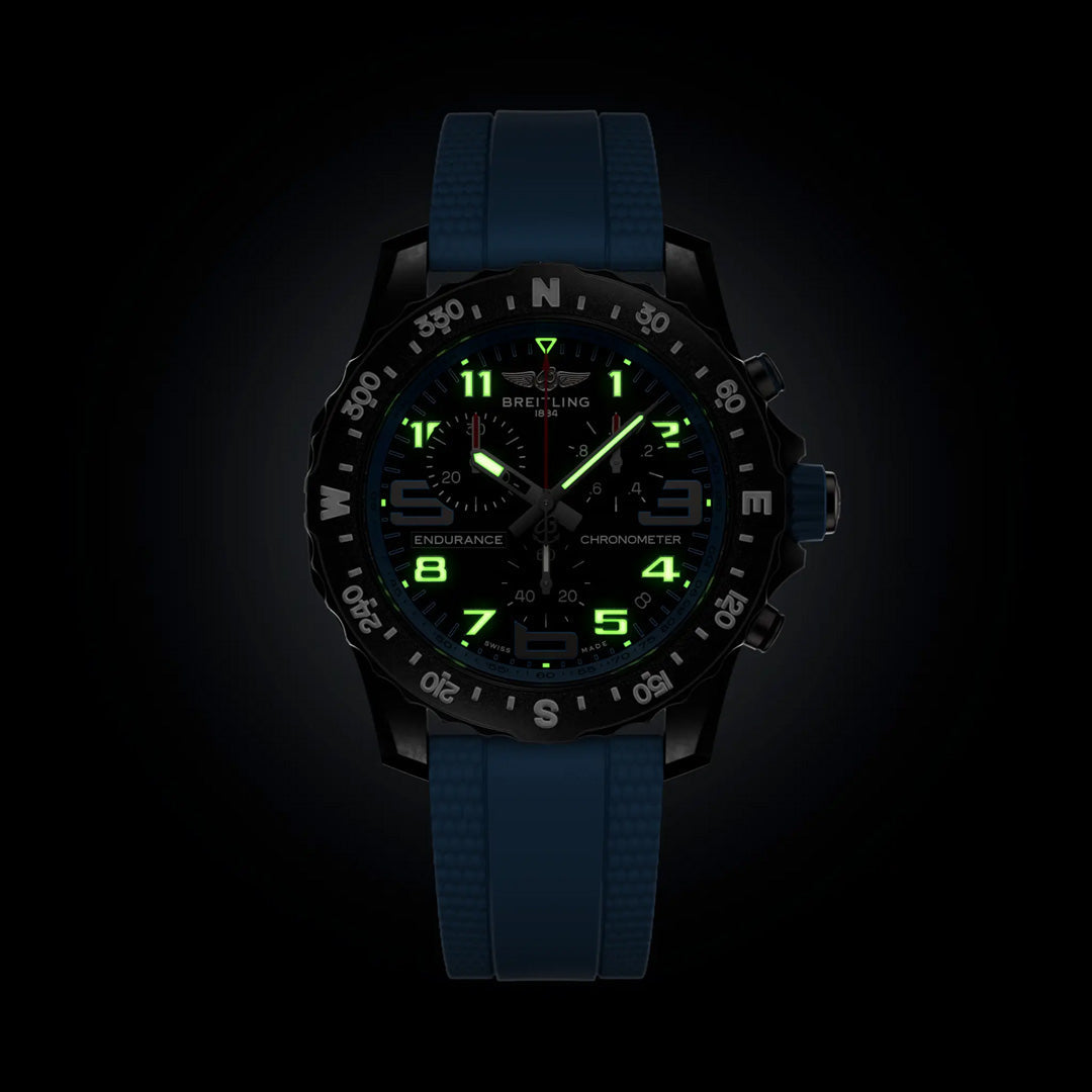 The BREITLING Professional Endurance Pro 44 features a blue strap and black dial. In darkness, its face and hands glow green, clearly illuminating the numerals and compass bezel for easy visibility.
