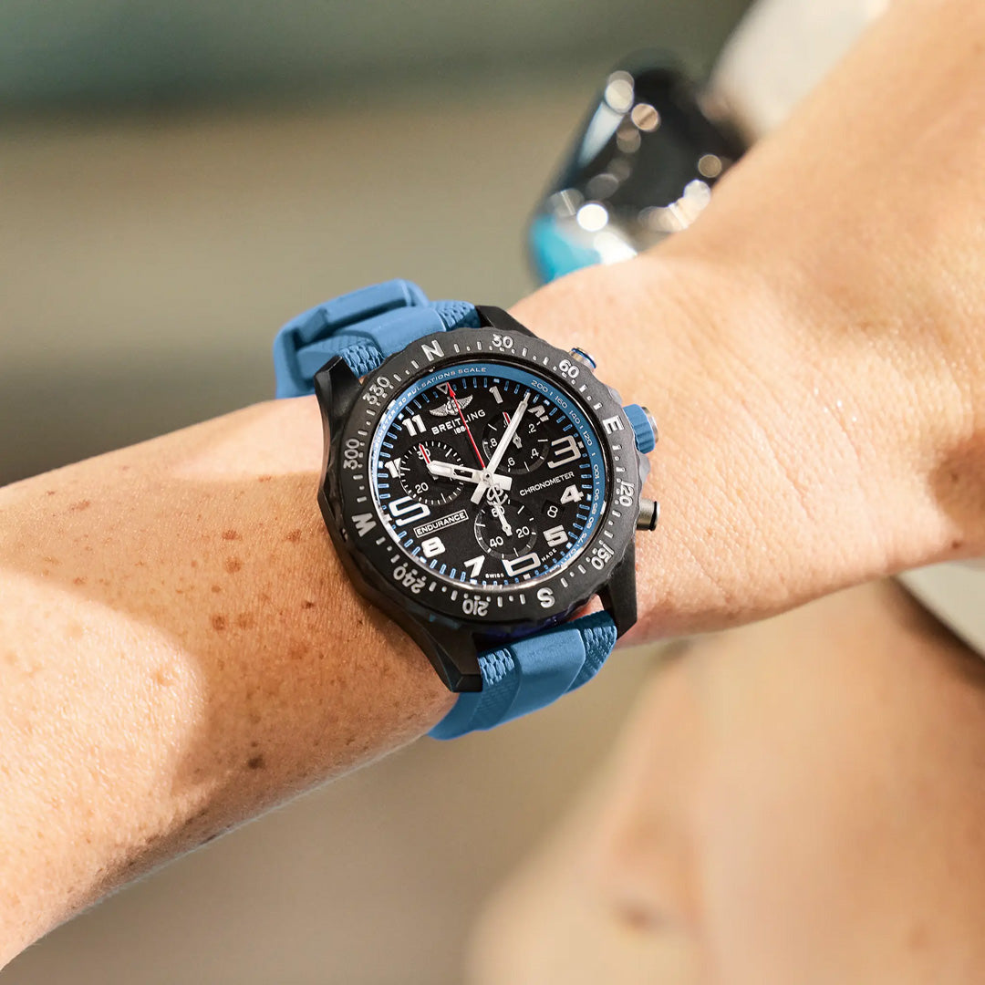 A close-up of a wrist wearing the Breitling Professional Endurance Pro 44 shows its sporty black face, blue strap, and distinctive dials and markers from Breitling, with the background artistically blurred.