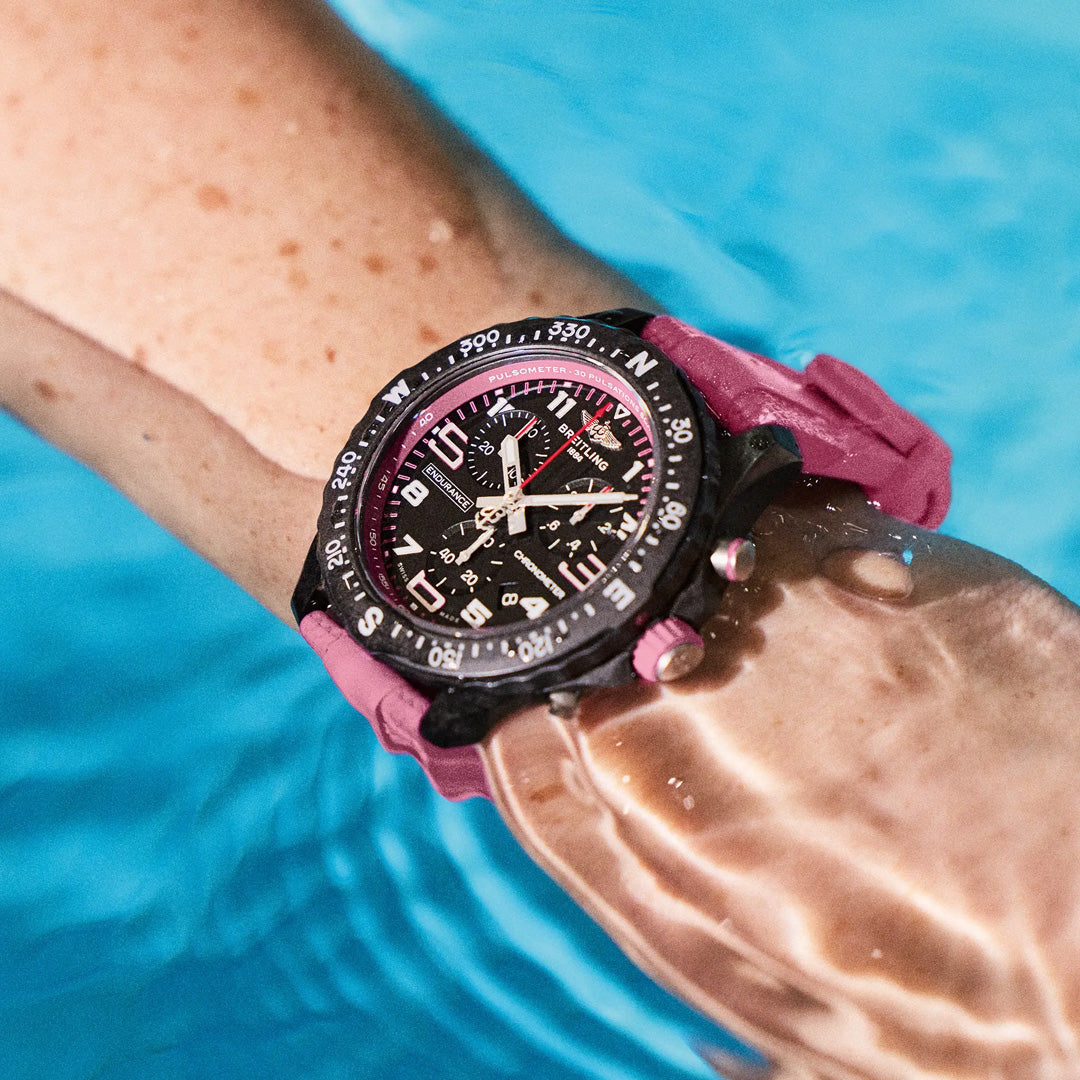 A close-up shows the BREITLING Endurance Pro 38 with a pink strap submerged in a pool. The watch features a black dial with white and red markings, compass bezel, and durable Breitlight® case. Water droplets on the watch and hand highlight its resilient design.