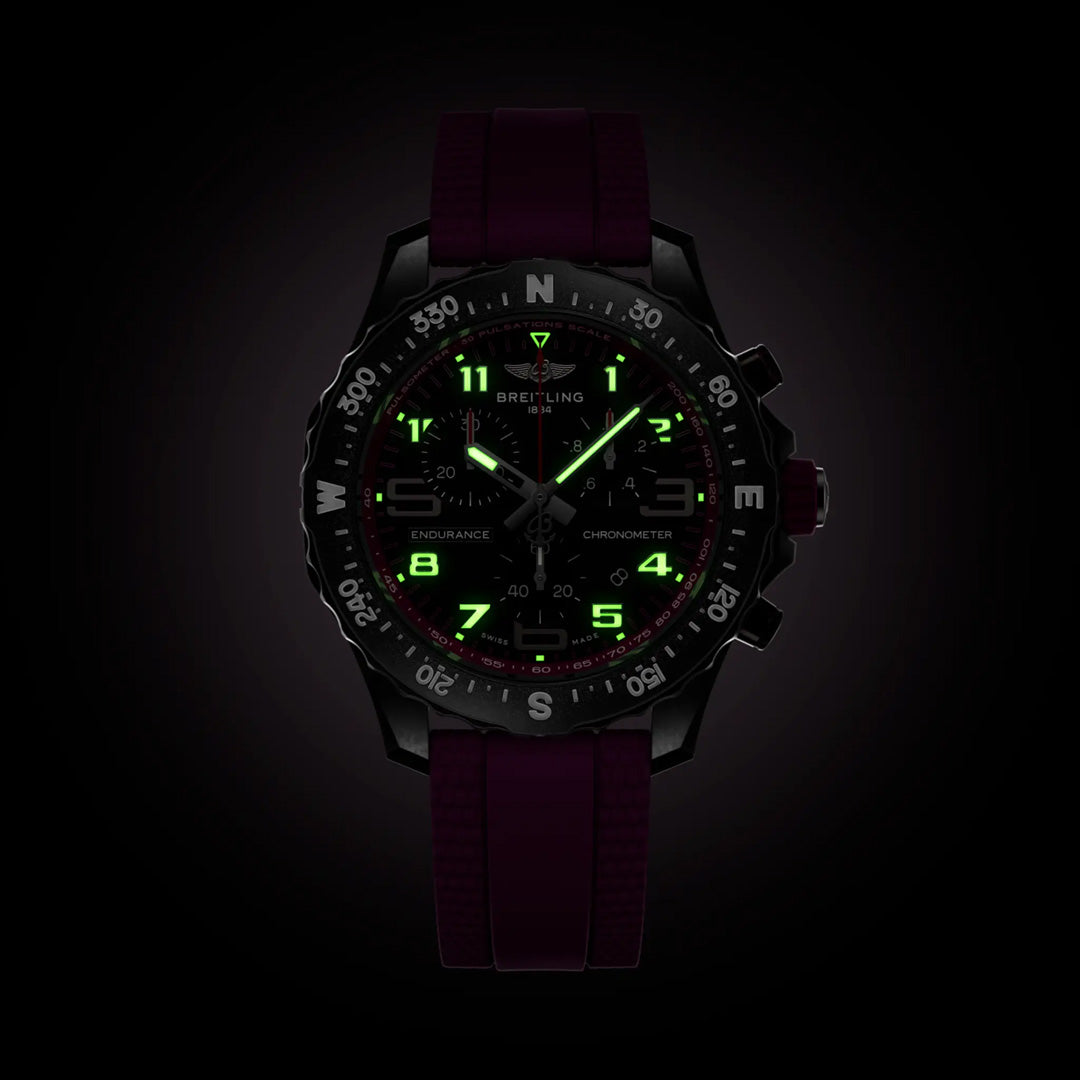 The BREITLING Endurance Pro 38 is a dark athleisure watch with a maroon strap, glowing green numerals, and hands. It features a black bezel with compass markings. The durable Breitlight® watch face displays the brand name and various dials highlighted against the dark background for visibility.