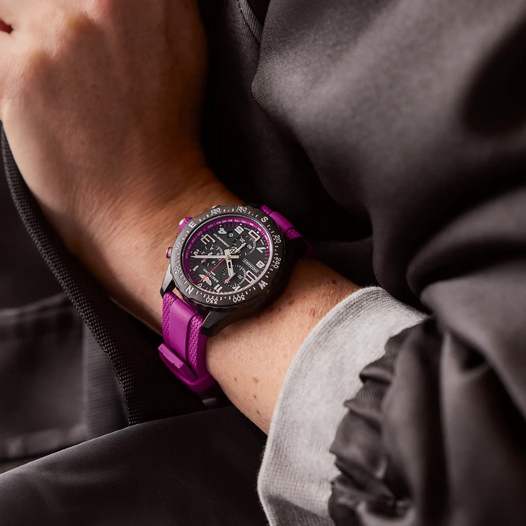 A person wearing a Breitling Endurance Pro 38, an athleisure watch with a purple strap. Its detailed black and white dial features various markers and a rotating bezel. Styled with a dark jacket, the gray sleeve beneath adds contrast to the stylish Breitlight timepiece.