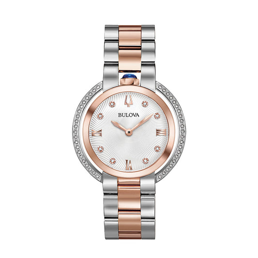 The Bulova Rubaiyat Quartz 35mm Watch features a stainless steel bracelet enhanced with elegant rose gold-tone accents. It has a round silver-toned dial with rose gold hour markers, while the bezel is beautifully decorated with small crystals, and the crown displays a distinctive blue accent.